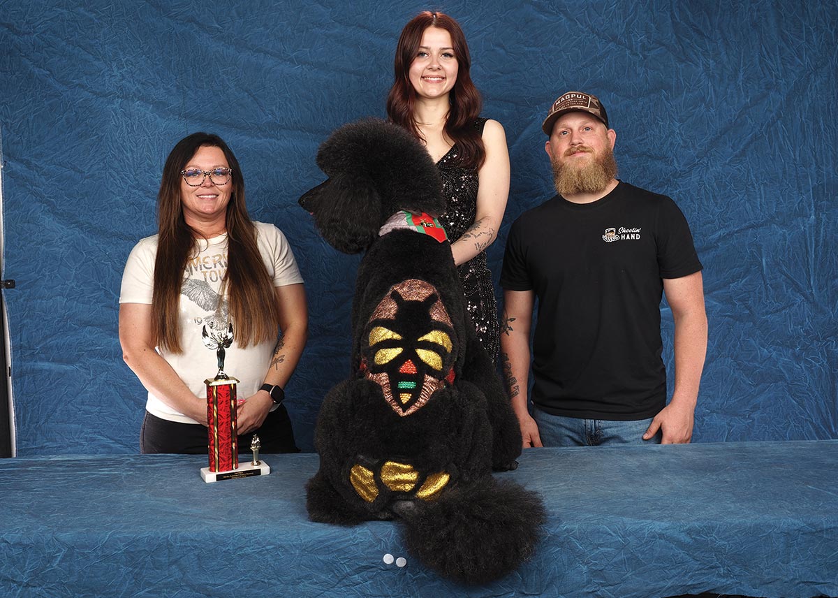 Three people pose with a black Poodle featuring creative coat carving and colorful dye on its back, next to a trophy.