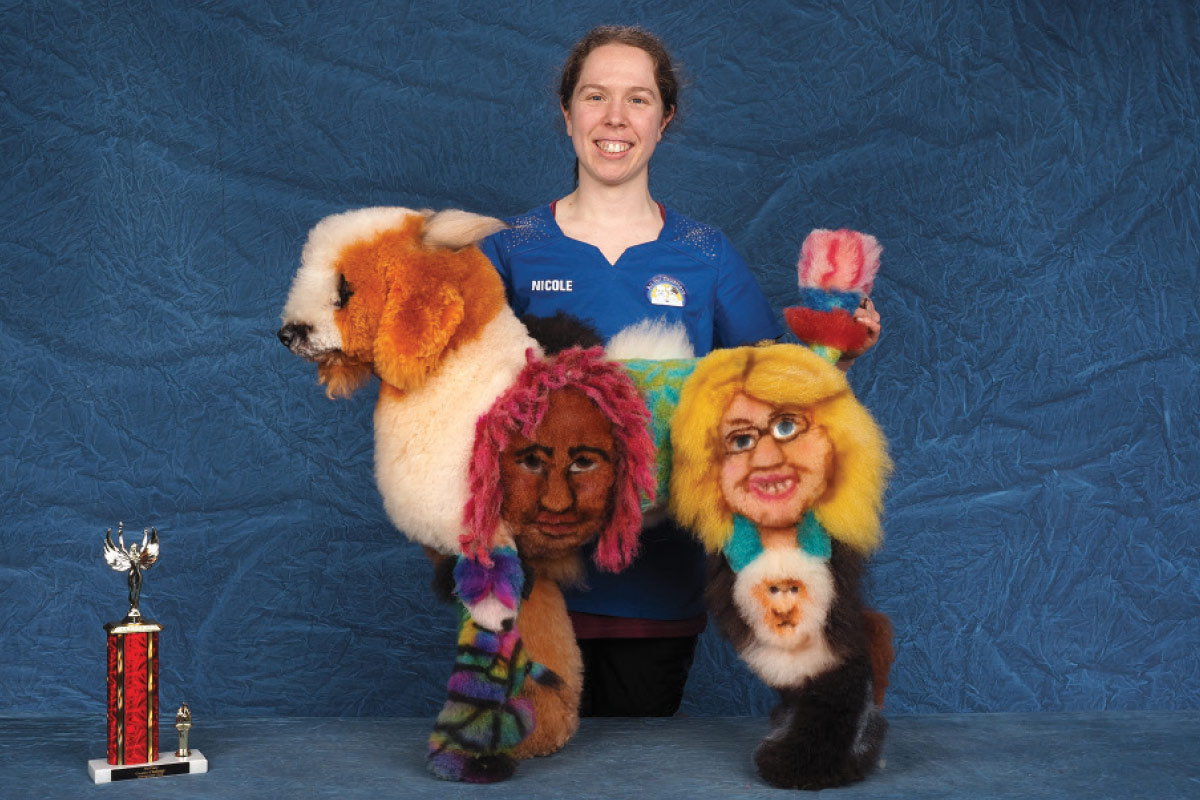 Woman poses with a Poodle creatively groomed and dyed with detailed portraits on its coat, shown next to a trophy.