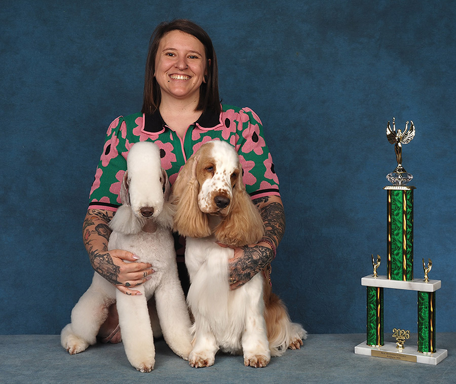 A smiling woman with tattooed arms poses with a Bedlington Terrier and a Cocker Spaniel alongside a green trophy.