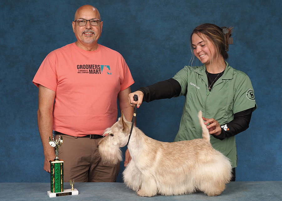 A groomer in a green smock poses a light-colored Scottish Terrier next to a presenter and trophy.