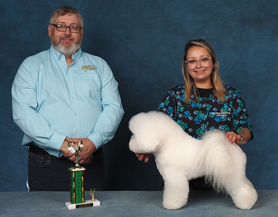 A groomer in a patterned smock holds a fluffy white Bichon Frise posed next to a presenter and trophy.