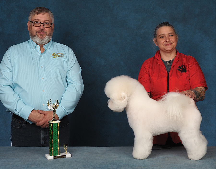 A woman in a red jacket poses a groomed white Bichon Frise beside a presenter and trophy.