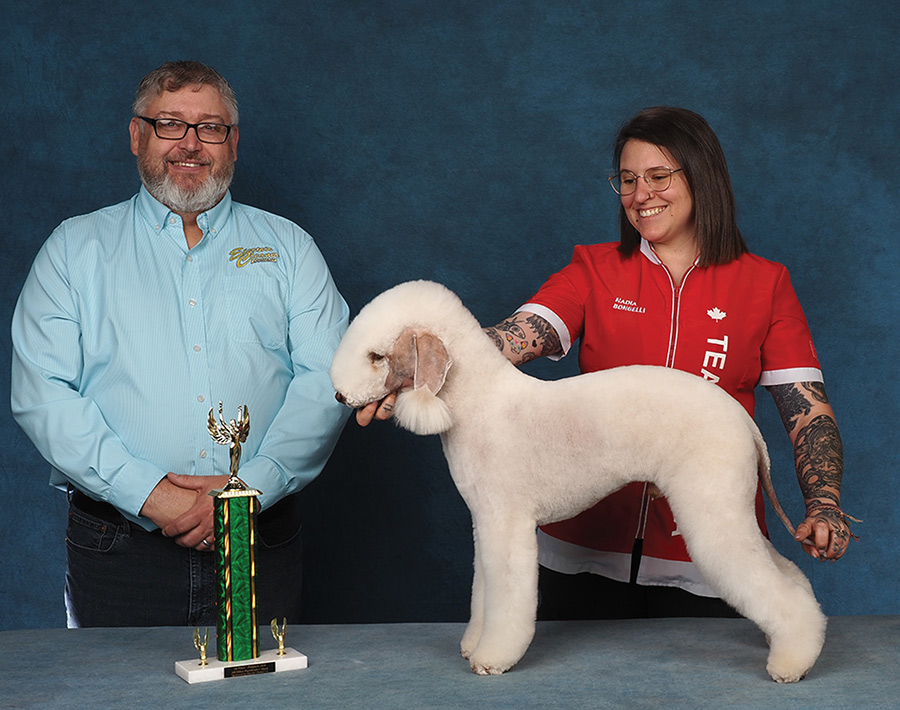 A woman in a red Team Canada jacket holds a groomed Bedlington Terrier posed next to a presenter and trophy.
