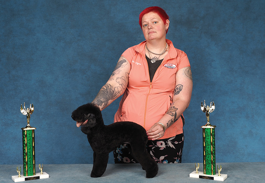 A woman with red hair poses a small black Poodle between two trophies.