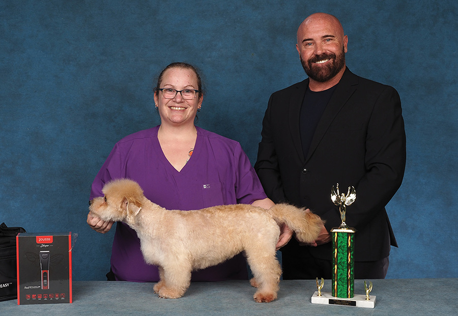 A groomer and presenter pose a tan dog next to grooming products and a trophy.