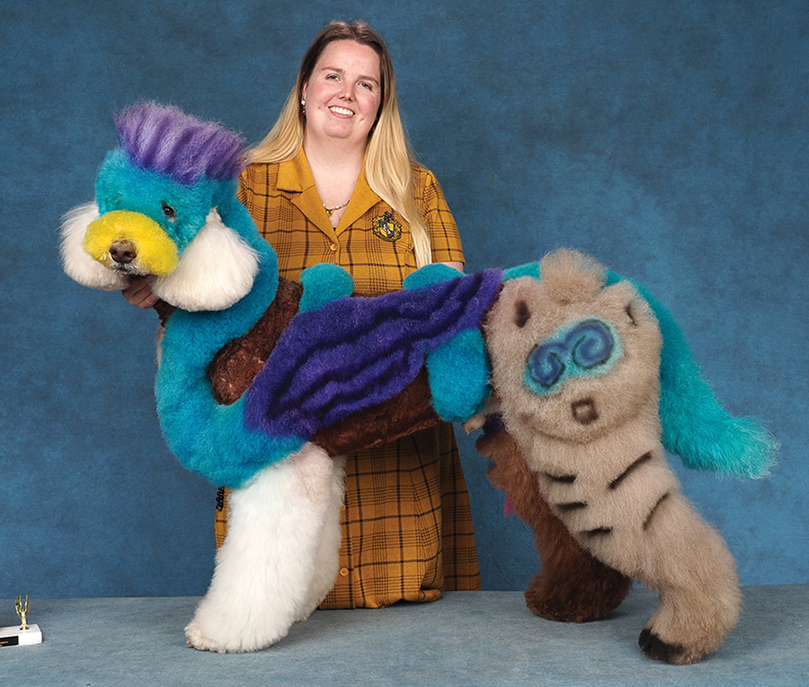 A groomer in a plaid outfit poses a Standard Poodle creatively dyed and styled with colorful cartoon designs.
