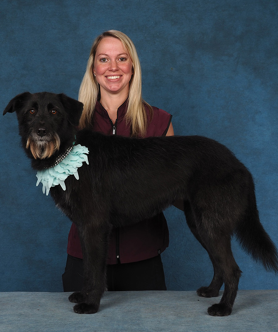 A smiling woman in a vest stands with a large dark dog adorned with a ruffled light blue collar.