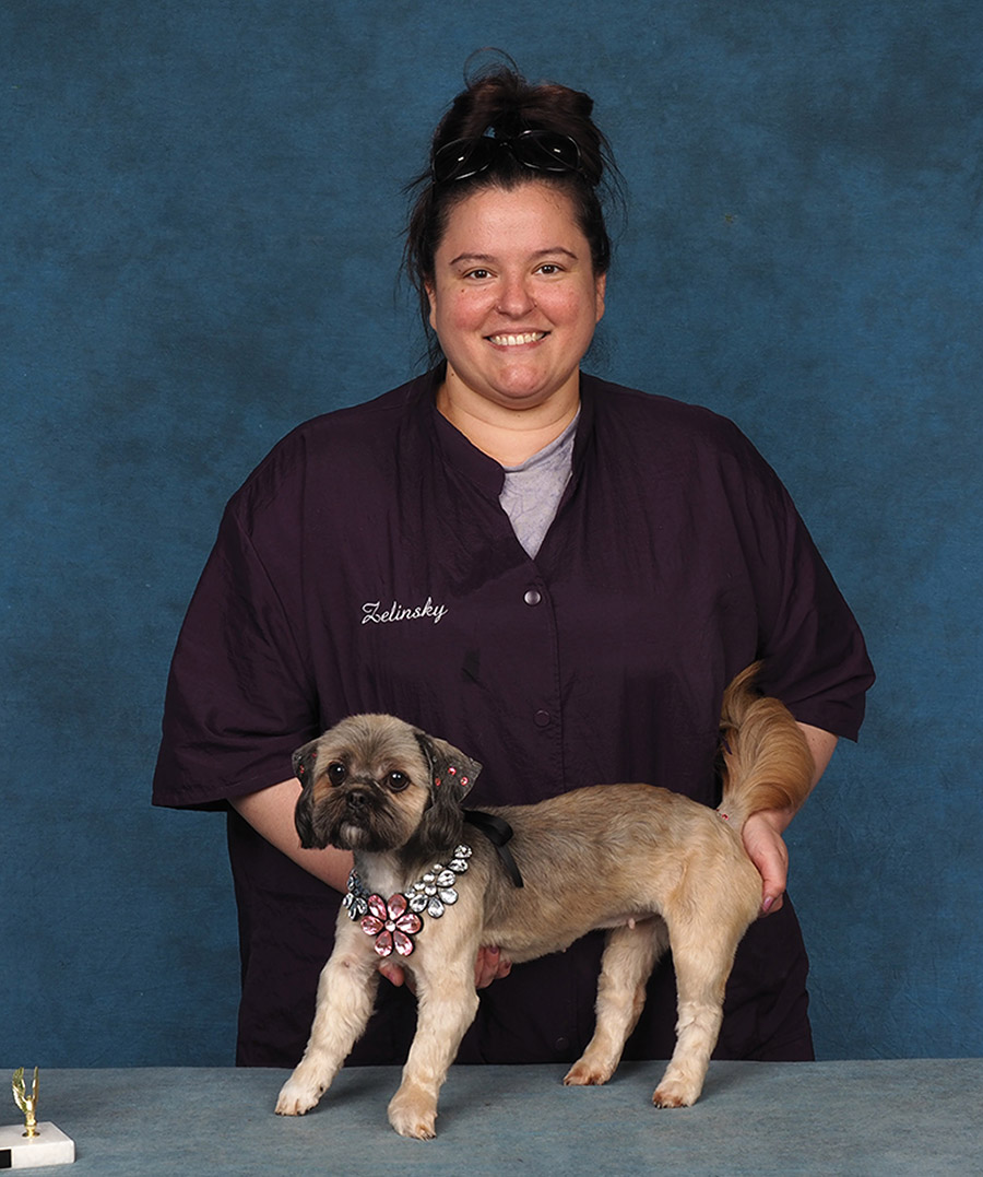 A smiling woman in a dark purple smock poses a small, light-colored dog adorned with a floral jewel necklace.