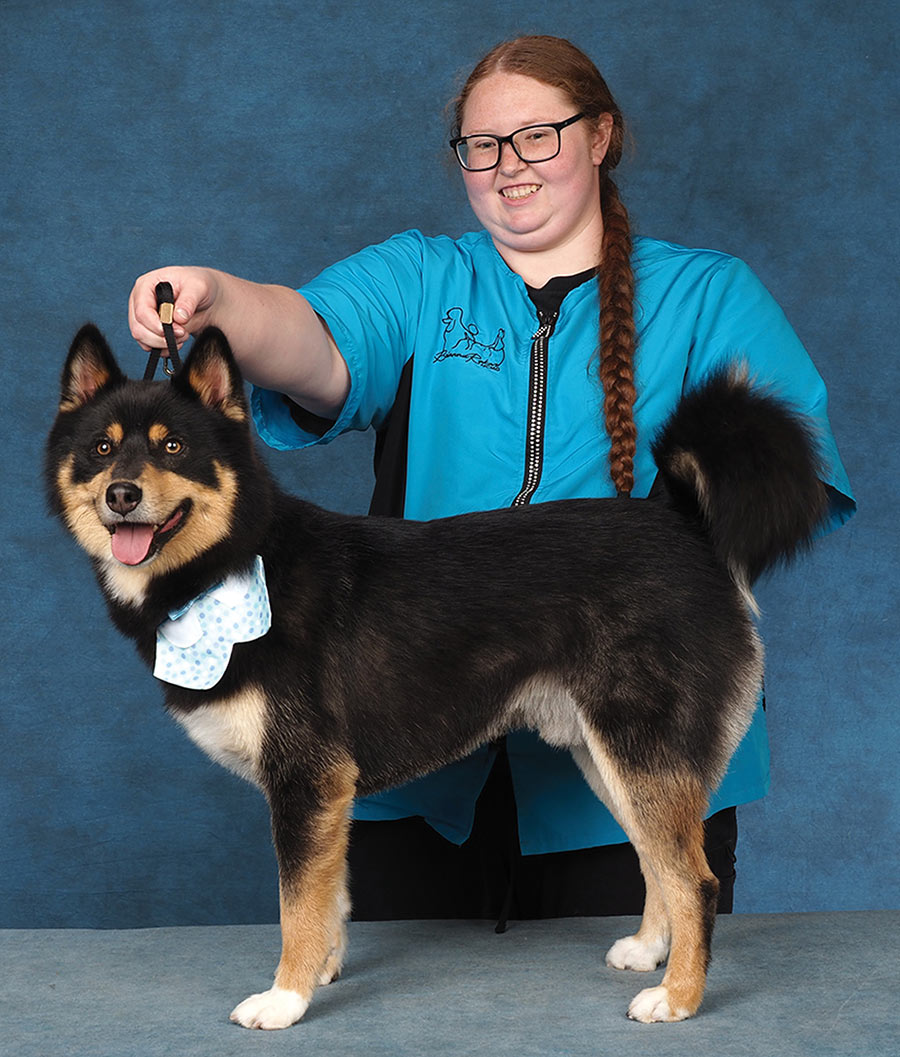 A smiling woman in a blue smock holds the leash of a black-and-tan Finnish Lapphund with a blue neckband.