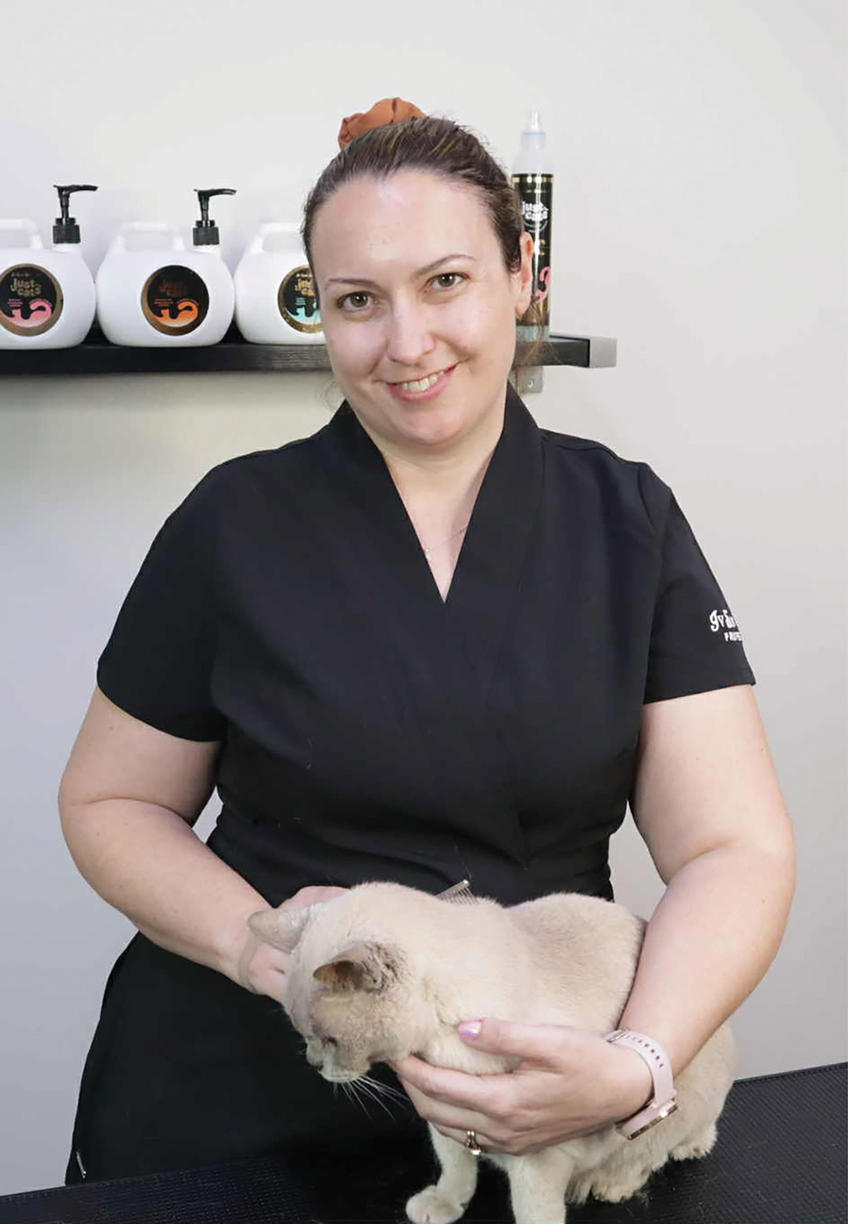 Smiling groomer holding a cream-colored cat at a salon table with product bottles behind.