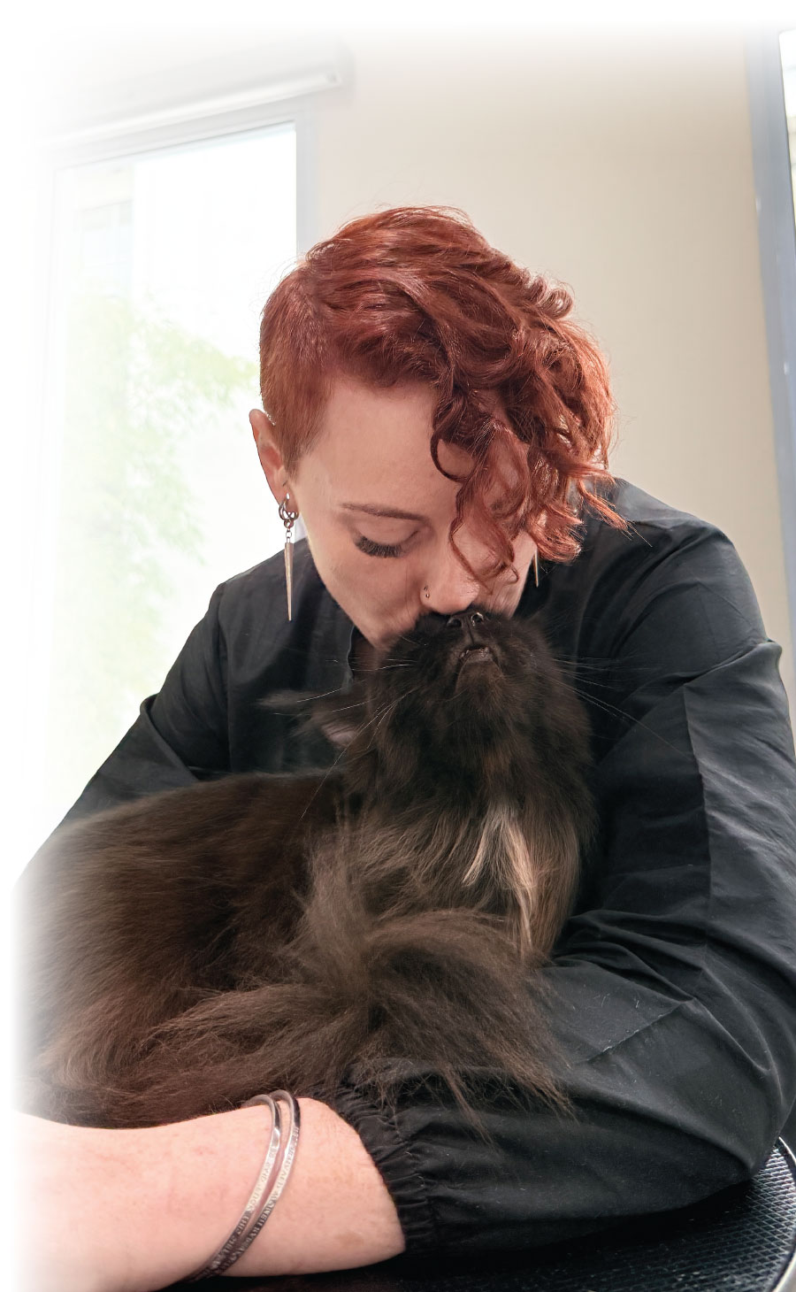 A groomer with short, curly red hair gently kisses the nose of a fluffy, long-haired dark cat she is holding in her arms.