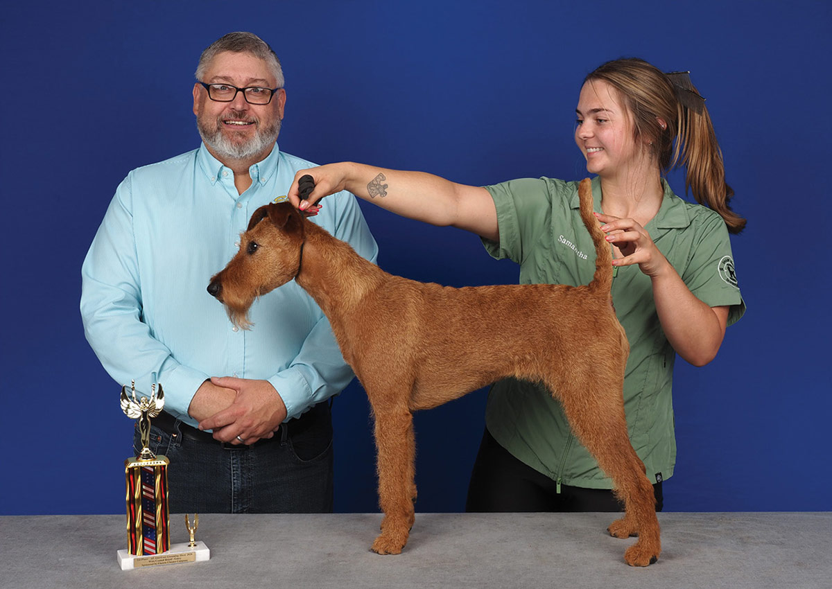 A man and a young woman in a green shirt hold a groomed Irish Terrier in a show pose with a small trophy, against a blue background.