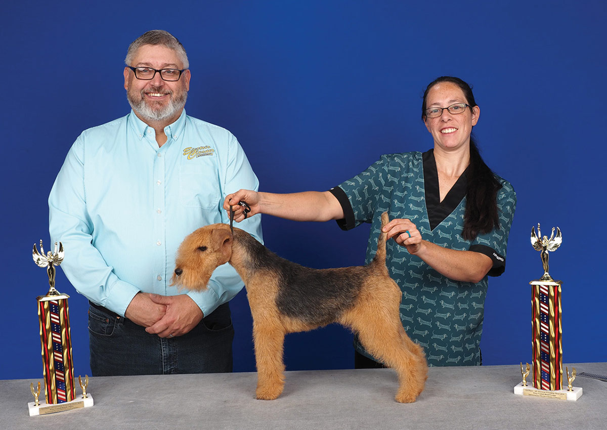 A man and a woman in a patterned scrub top pose with a groomed black and tan Welsh Terrier in a show stance, with two small trophies, against a blue background.