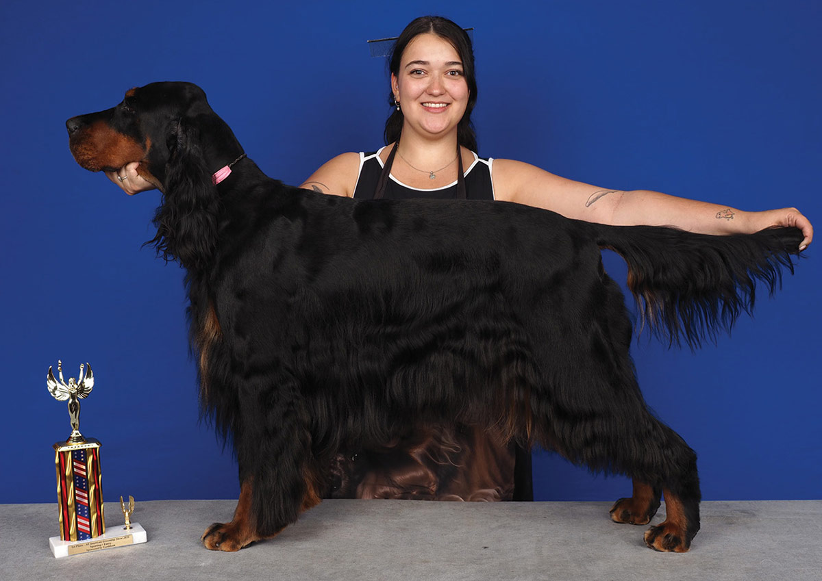 A woman smiles as she poses with her well-groomed, large black and tan dog beside a tall trophy, all set against a solid blue background.