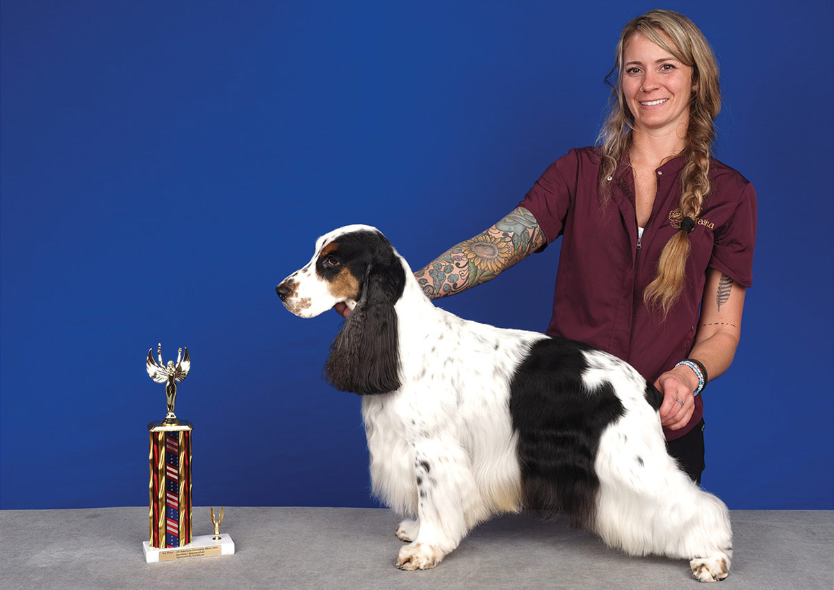 A tattooed woman smiles while presenting her groomed parti-colored black and white spaniel beside a gold trophy against a blue background.