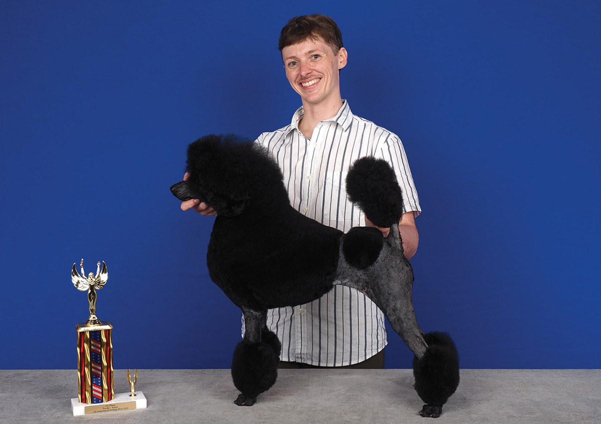 A smiling man poses a black poodle in a show clip beside a trophy on a blue background.