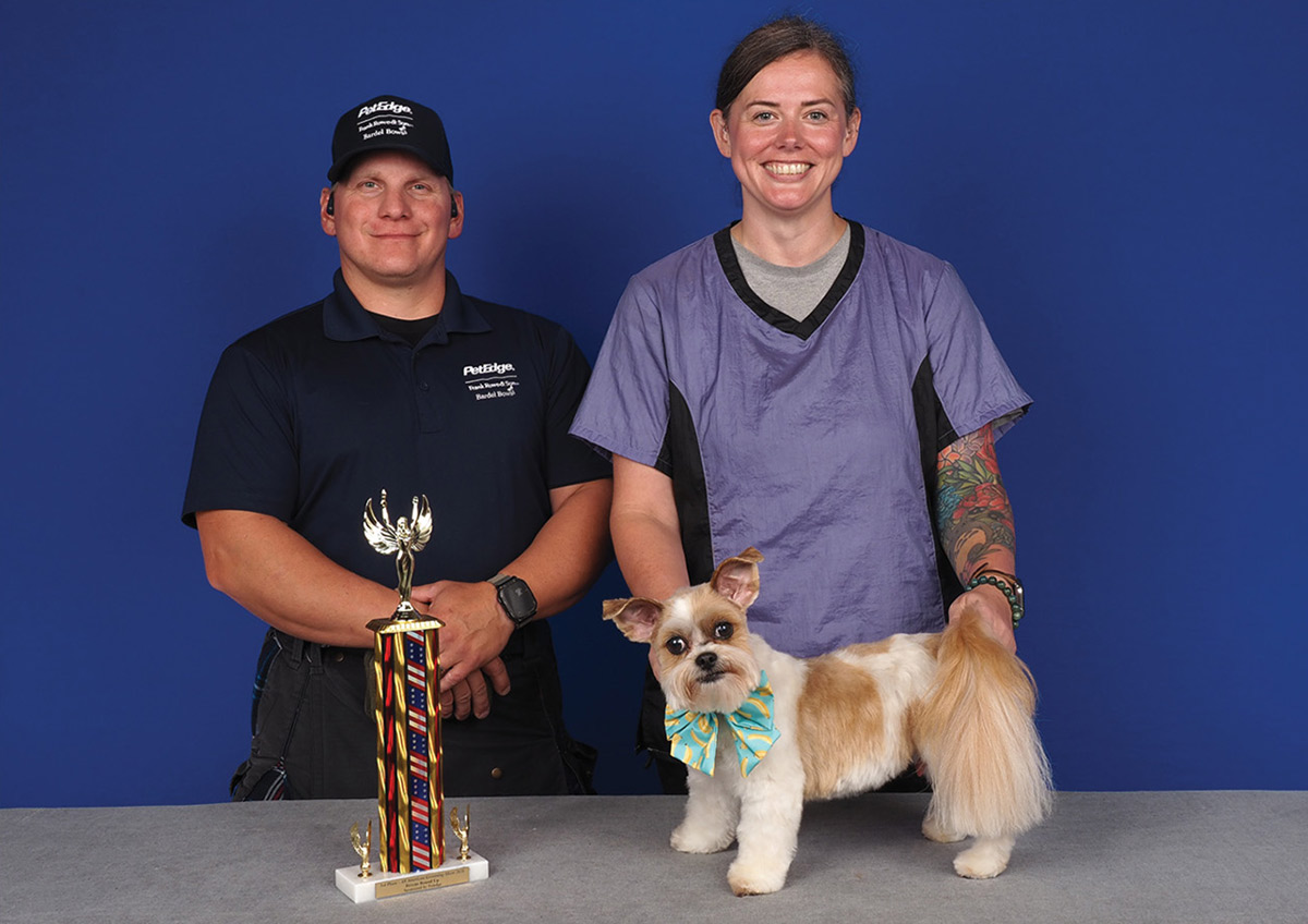 A dog groomer and an official stand behind a small white and tan dog wearing a patterned bowtie on a table beside a tall trophy.