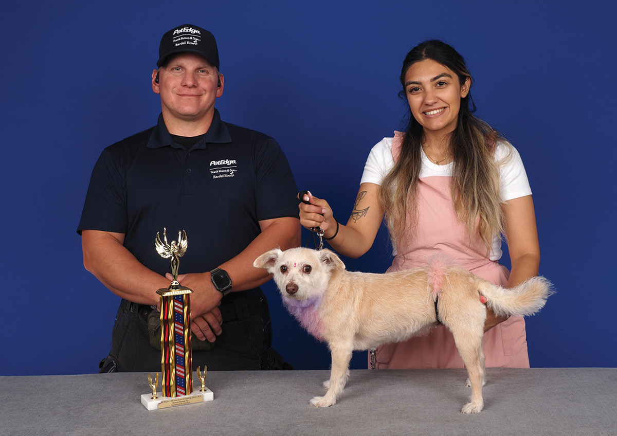 A dog groomer holds a small light-tan dog with pink accent dye next to an official and a trophy at a pet grooming competition.