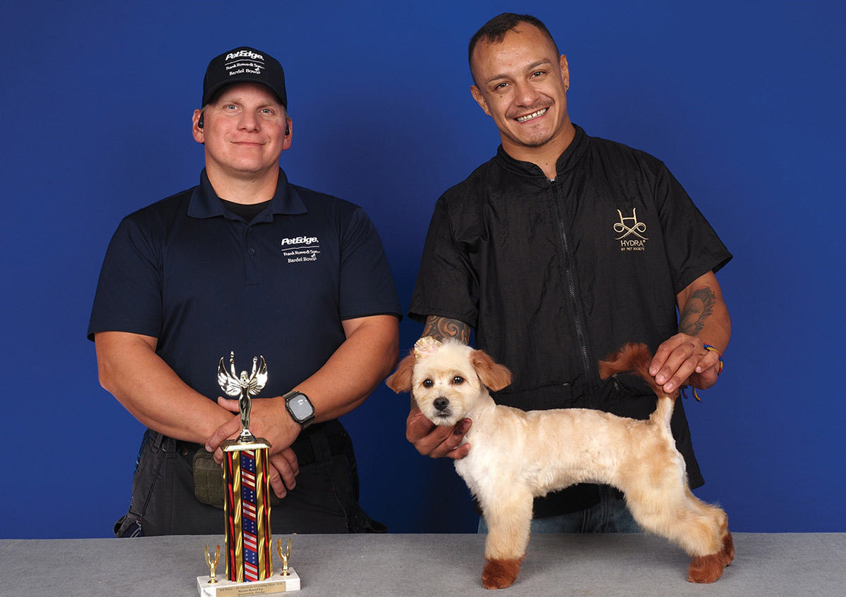 A dog groomer holds a small tan dog with dyed reddish accents and an ear bow, posing beside a competition official and a tall trophy.