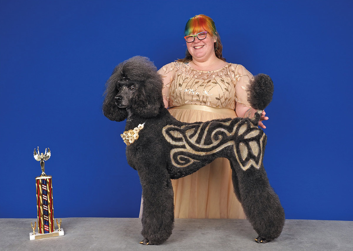 A smiling groomer stands behind a black poodle with intricate gold glitter patterns clipped into its coat, posing on a table next to a trophy.