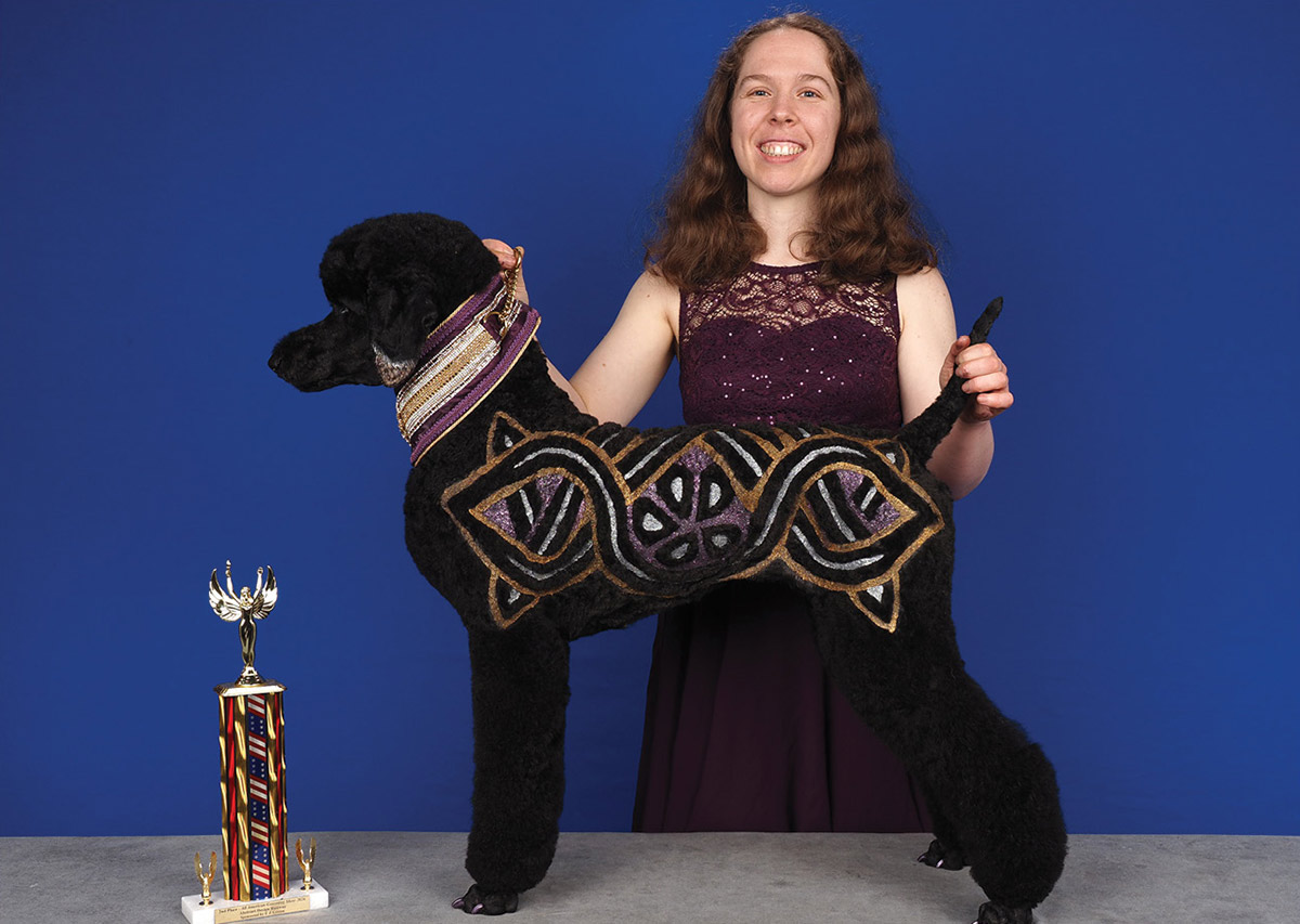 A smiling groomer poses behind a standing black poodle styled with intricate purple, gold, and silver geometric fur designs and a decorative collar next to a trophy.