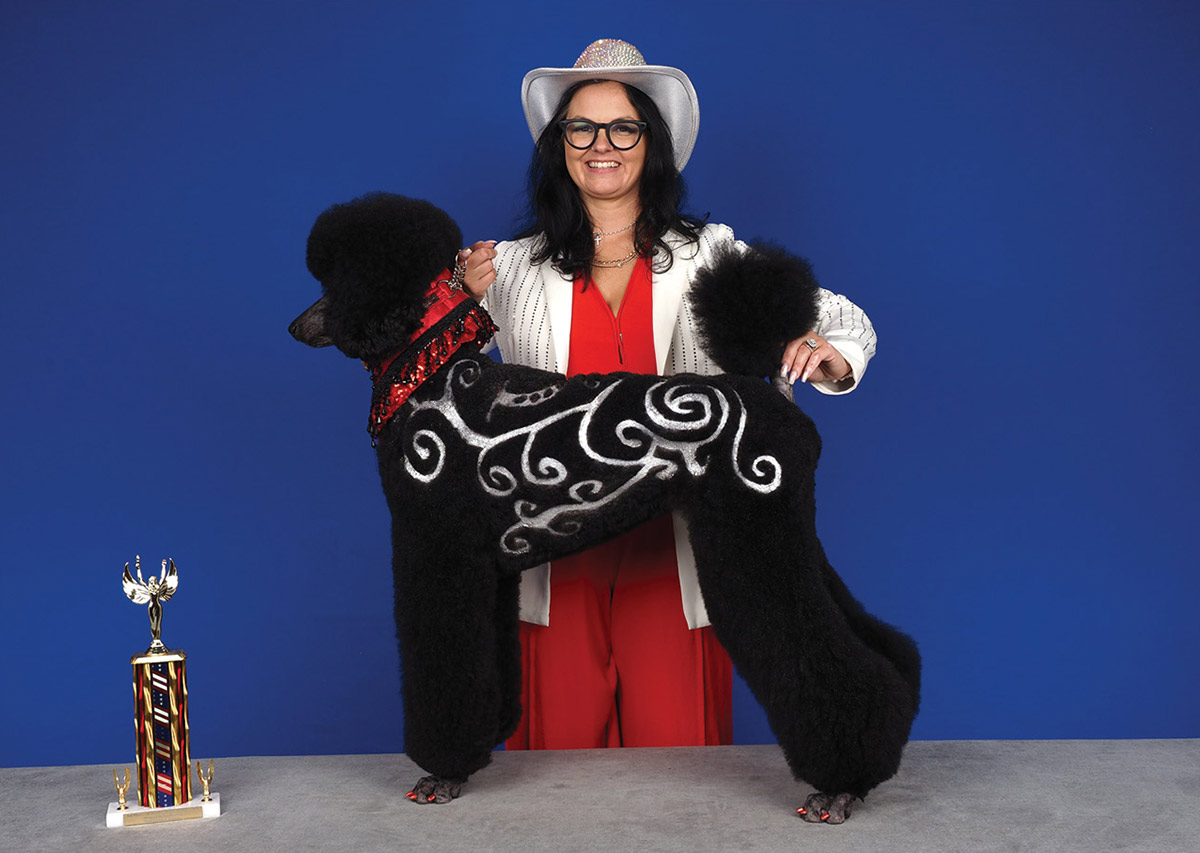 A smiling groomer in a cowboy hat poses behind a standing black poodle styled with silver glitter scroll designs and a red sequined collar, beside a trophy.