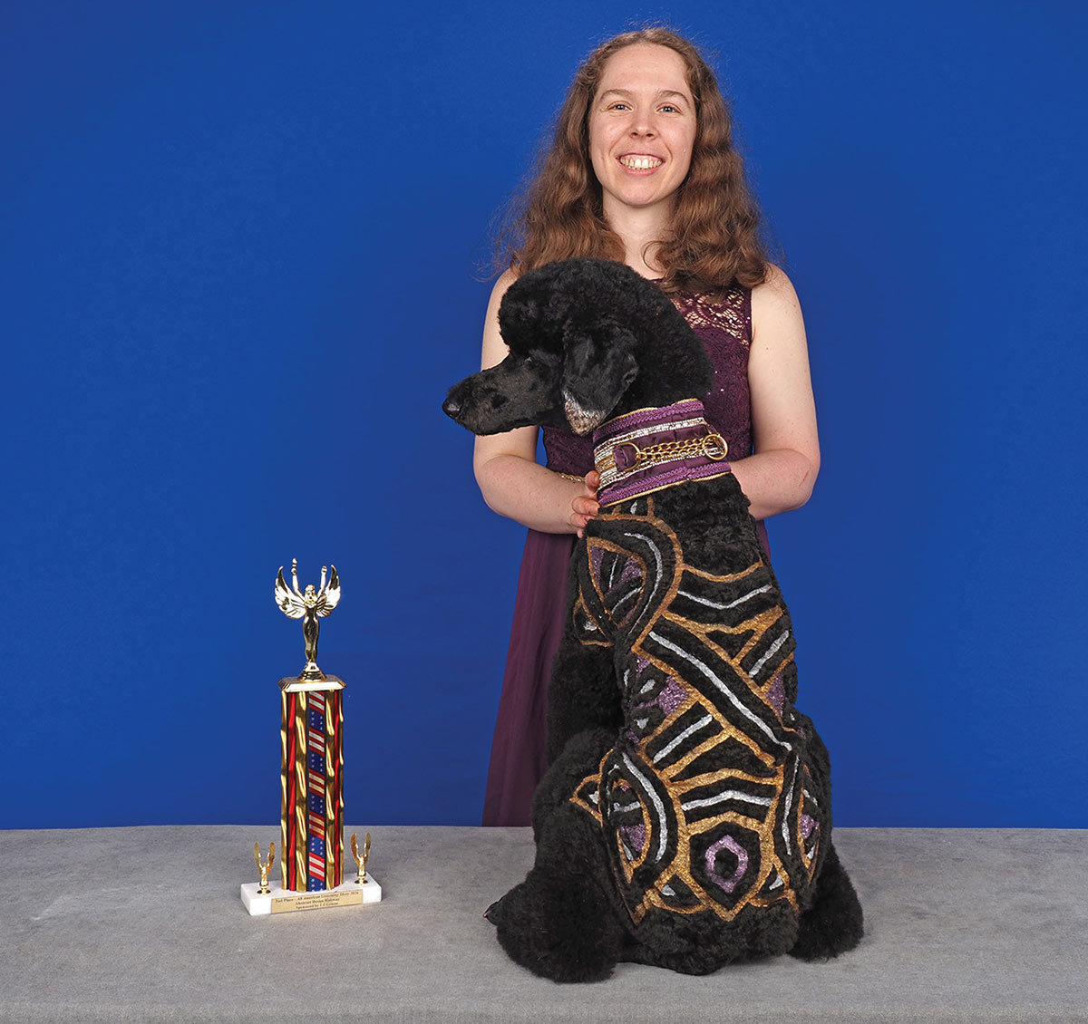 A groomer poses behind a sitting black poodle, showing off ornate purple, gold, and silver clipped patterns running down its back next to a competition trophy.