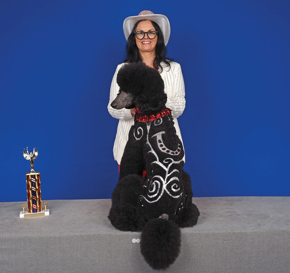 A groomer in a cowboy hat stands behind a sitting black poodle, showcasing silver glitter swirl and horseshoe patterns etched into its back next to a trophy.