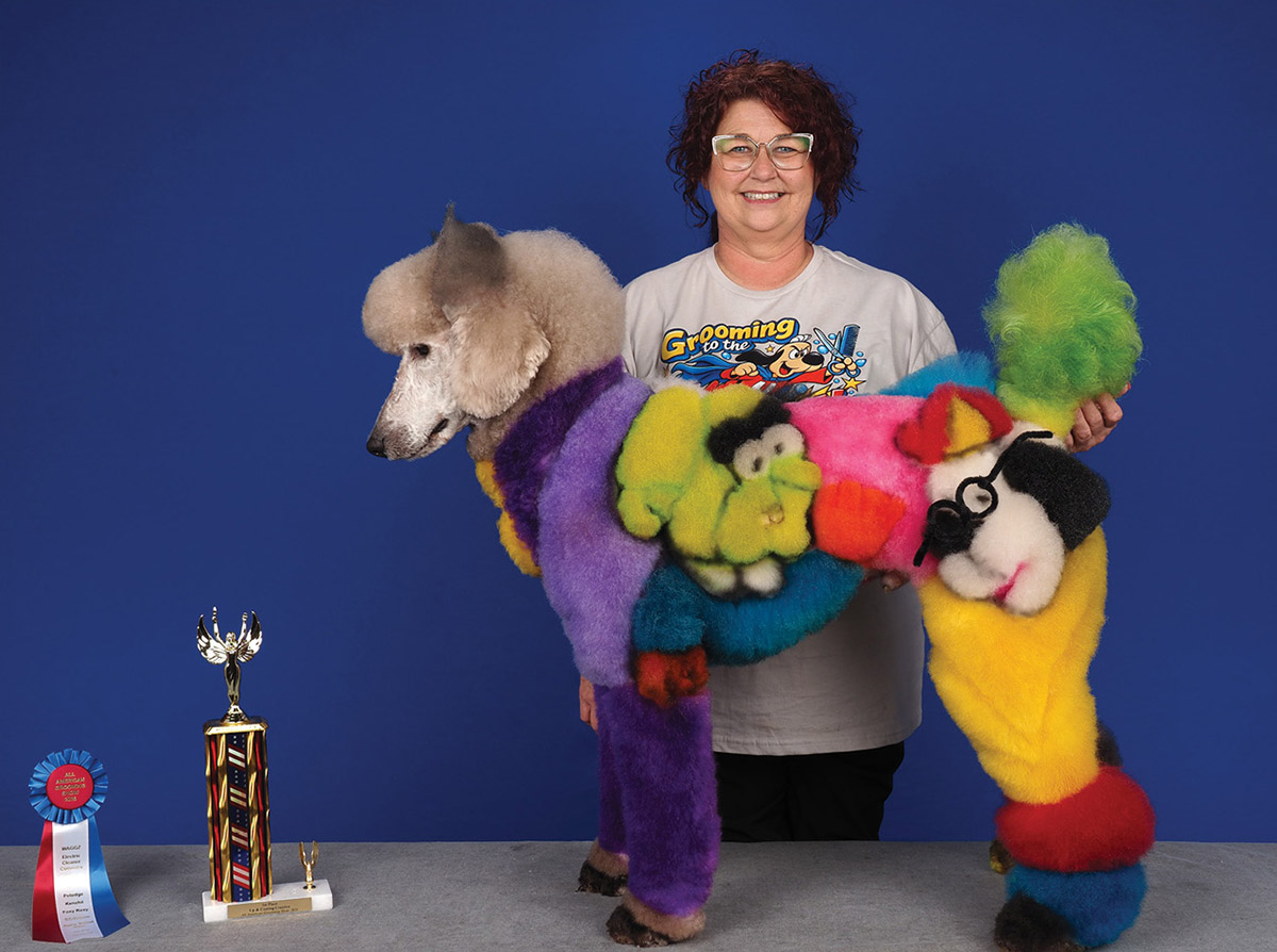A smiling groomer poses behind a standard poodle creatively dyed with vibrant cartoon character designs, standing next to a trophy and award ribbon.