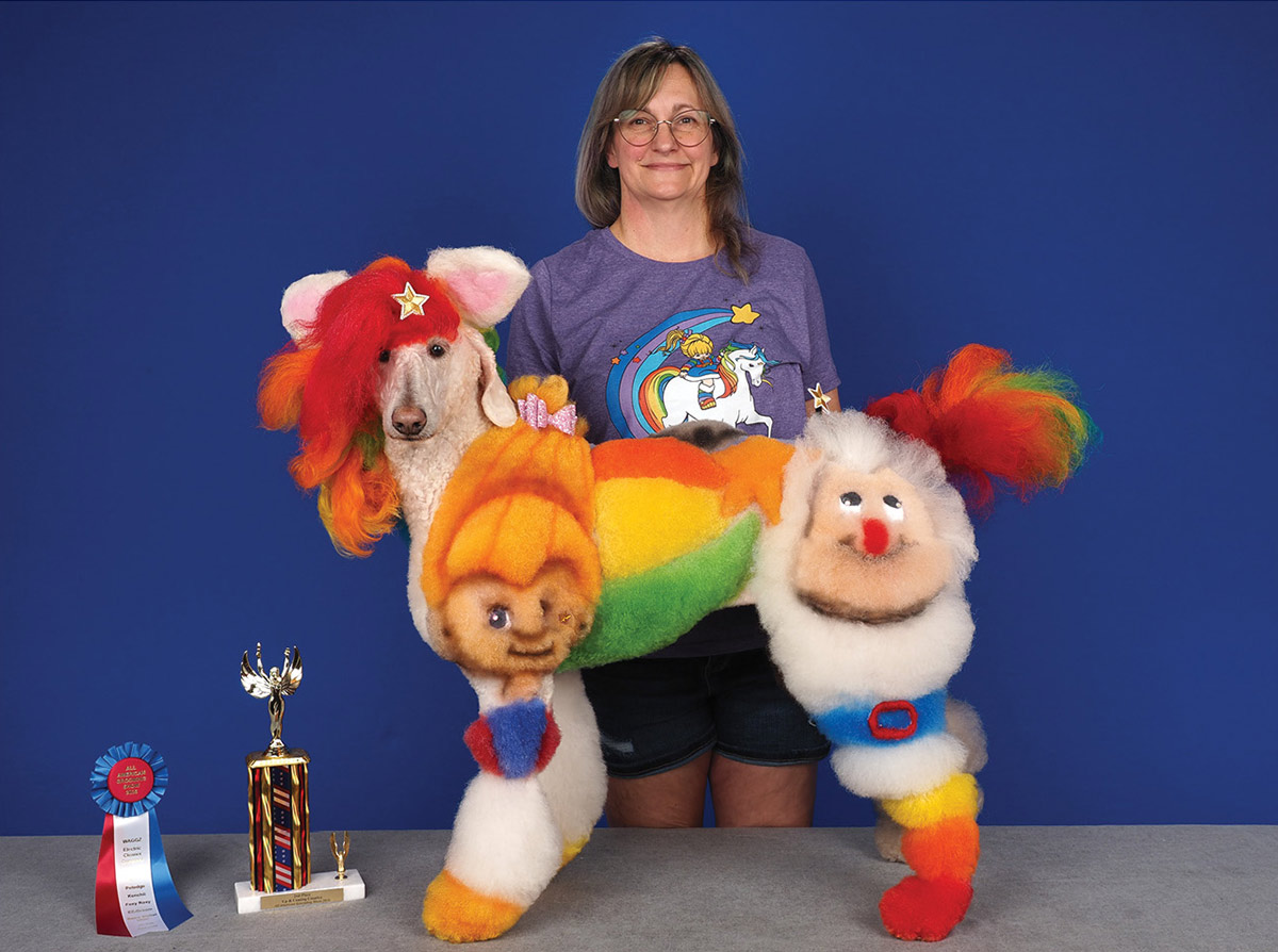A groomer poses behind a standard poodle creatively dyed with bright Rainbow Brite characters and rainbow fur, standing next to a competition trophy and award ribbon.