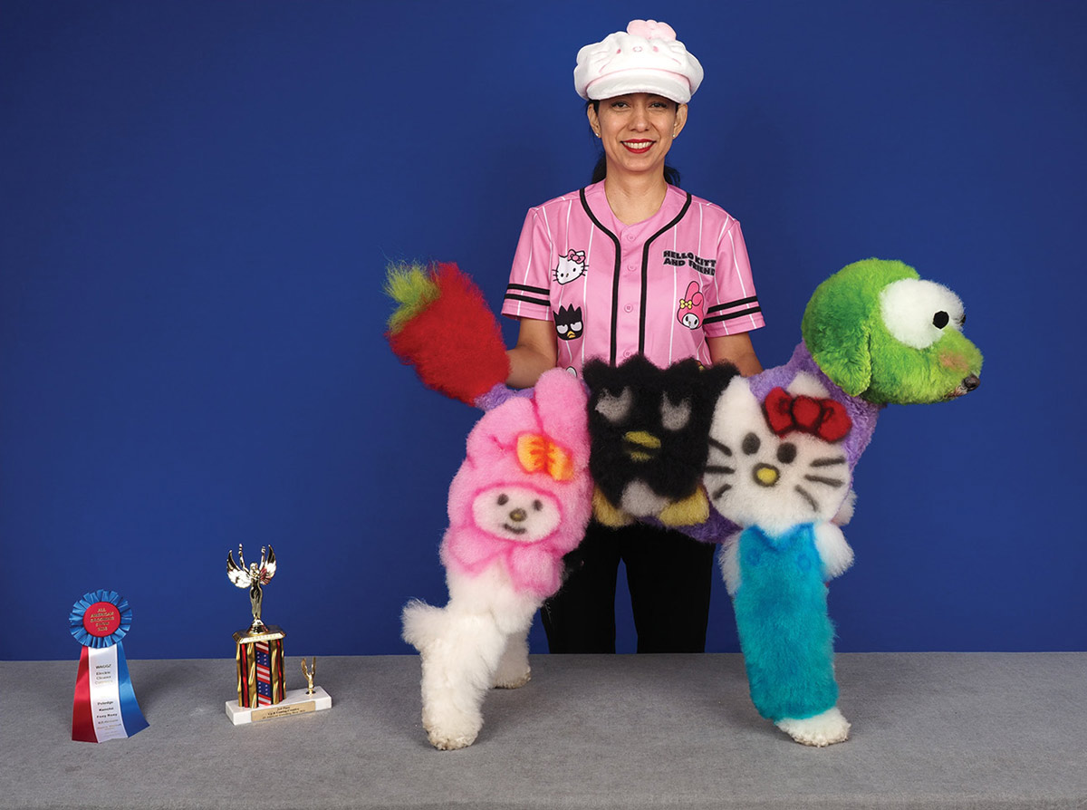 A groomer in a pink shirt poses behind a poodle creatively dyed with colorful Hello Kitty and Sanrio character designs next to a trophy and award ribbon.