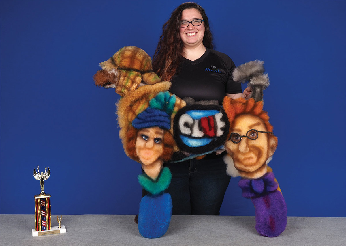 A smiling groomer poses behind a poodle creatively dyed with the Clue board game logo and character portraits, standing next to a competition trophy.