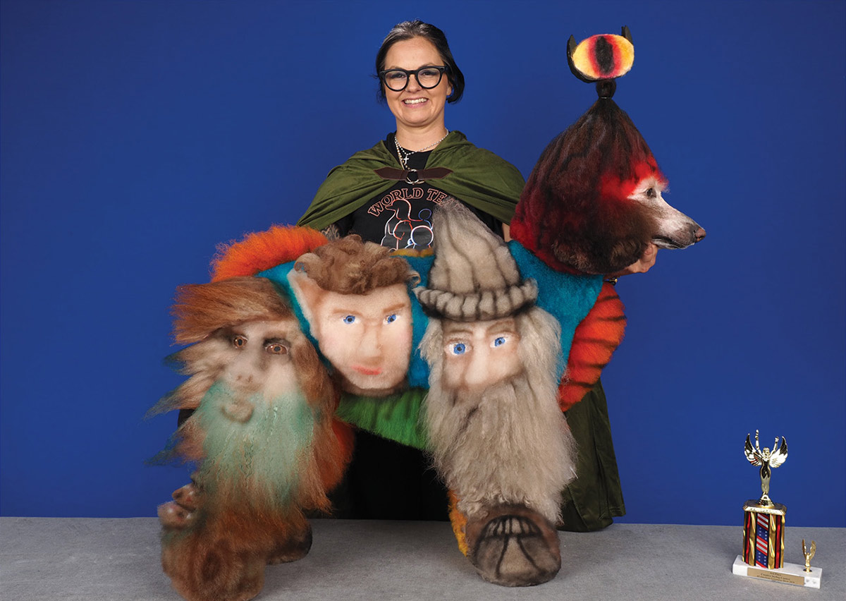 A groomer in a green cape poses behind a poodle creatively dyed with Lord of the Rings characters, including Gandalf and the Eye of Sauron, standing next to a competition trophy.