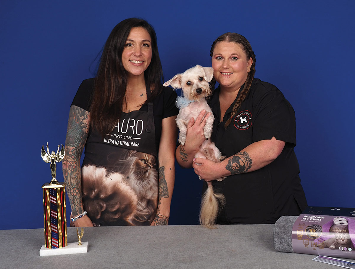 Two women pose behind a grooming table, one holding a small light-colored dog with a blue bow, standing next to a competition trophy.