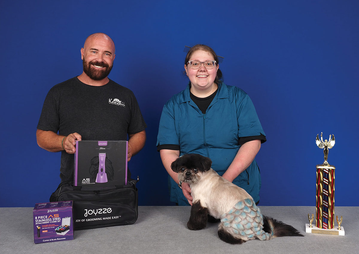 A groomer and presenter holding Joyzze products pose with a small dog featuring teal scale-patterned fur dye, standing next to a competition trophy.