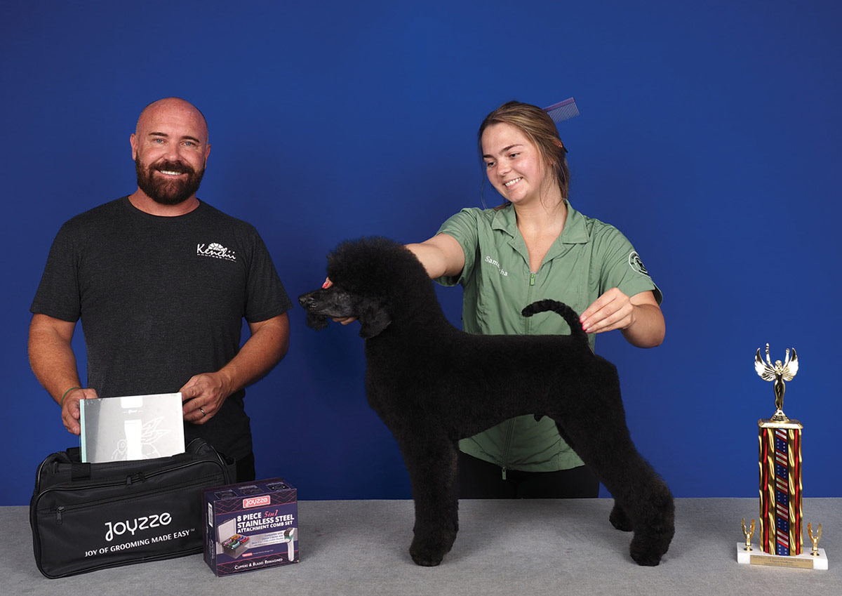 A groomer stacks a freshly trimmed black poodle on a table next to a presenter holding Joyzze products and a tall competition trophy.