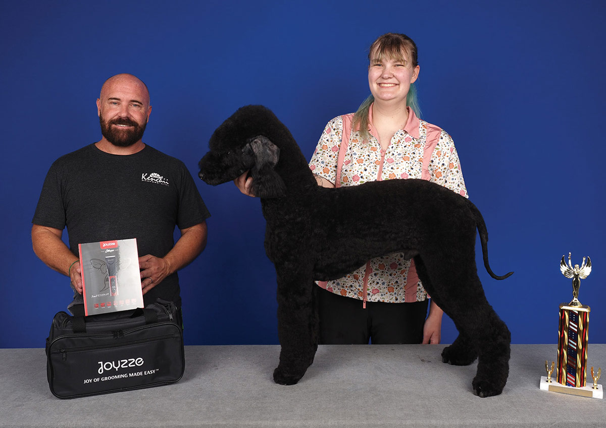 A groomer poses with a large black poodle next to a presenter holding Joyzze clippers and a tall grooming competition trophy.
